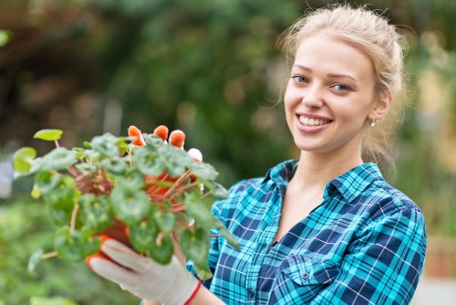 A young woman with blonde hair tied back, smiling while holding a small potted plant with green, rounded leaves and pinkish-red flowers, in an outdoor garden setting. Behind her, there are lush, green shrubs and trees, indicating a well-maintained backyard garden in Leyton. She is dressed casually in a blue plaid shirt and wearing gardening gloves, suggesting she is engaged in gardening activities. The background shows a variety of natural plant life with dense foliage, and the lighting indicates a bright day with soft shadows, typical of mild weather. The scene emphasizes garden care and plant nurturing, aligning with outdoor gardening and landscaping services offered by local providers such as Landscapers Leyton.