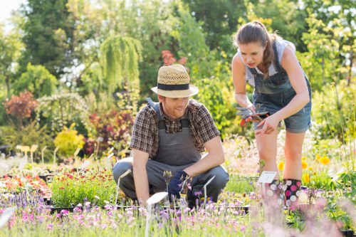 A brightly lit outdoor garden scene showing two women working together in a landscaped yard, with one crouching close to the ground tending to flower beds containing a variety of flowering plants in shades of pink, purple, and yellow. The other woman is standing beside her, holding garden shears and wearing a denim dress over a white shirt. Behind them, there are lush green trees and shrubs providing a natural background, with some taller trees in the distance and a well-maintained lawn area in the foreground. The garden features a mix of flower beds bordered by wooden edging, a paved pathway, and patches of vibrant flower vegetation, indicative of a well-maintained outdoor space suitable for gardening and landscaping services by Landscapers Leyton, with natural sunlight enhancing the scene’s colours and textures.