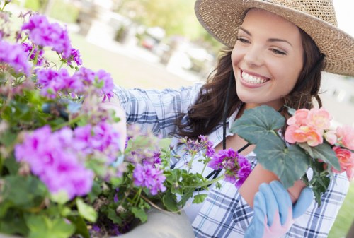 A woman with long brown hair, wearing a wide-brimmed straw hat, a blue and white checkered shirt, and light blue gardening gloves, is smiling as she tends to vibrant flowering plants in a garden in Leyton. She is holding a pink flower with large green leaves in one hand, and arranging purple and pink flowers in a white ceramic pot with the other. The garden features a lush green lawn, bordered by flower beds with a variety of colourful blossoms, and a background with trees and soft sunlight filtering through the leaves, creating a bright and cheerful outdoor setting suitable for landscaping and gardening care services in Leyton.