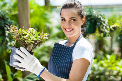 A woman with dark hair pulled back, wearing a white t-shirt, a blue striped apron, and white gardening gloves, stands in a lush garden with vibrant green foliage. She is holding a small potted shrub with green and reddish leaves, smiling at the camera. In the background, there are various plants, trees, and a wooden post, indicating a well-maintained outdoor space. The garden includes grass, flower beds, and paving areas, all showcasing healthy plant life. The scene is brightly lit with natural daylight, suggesting a clear, sunny day. This image visually relates to professional gardening and landscaping services, possibly provided by Landscapers Leyton, offering expertise in plant care, garden maintenance, and outdoor landscaping within Leyton or nearby areas in London.