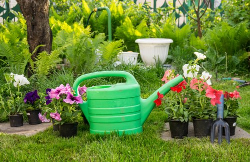 The image depicts a well-maintained garden area with a lush green lawn featuring neatly cut grass. In the foreground, there are multiple potted flowering plants with vibrant pink, purple, and white blooms, arranged along the edge of the lawn. A green plastic watering can is centrally positioned, surrounded by the potted plants, indicating active gardening. Behind the watering can, there are dense green ferns and other foliage, contributing to the garden's lush appearance. Further in the background, a large mature tree trunk is visible, providing shade, and additional garden elements like a white decorative pot and a garden fork are partially visible, suggesting ongoing landscaping or planting activities. The overall scene is outdoors on a clear day, with natural lighting highlighting the bright, natural tones of the plants and garden tools, creating a welcoming and tidy outdoor space suitable for gardening services provided by companies such as Landscapers Leyton.