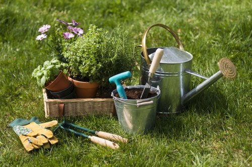 A well-maintained backyard garden features a lush, green lawn with evenly cut grass, bordered by a neat flower bed containing a variety of potted plants and flowers in shades of purple, white, and green. In the foreground, gardening tools including a pair of gloves, a small garden fork, and hand trowels are laid on the grass. To the right, there are a metal watering can with a wooden handle, a silver bucket filled with soil, and a small plastic trowel resting inside. The background reveals a mixture of mature trees and hedges, creating a private outdoor space suitable for gardening and outdoor activities. The scene is illuminated by natural daylight, suggesting a clear, bright weather condition typical of a spring or summer day in Leyton, east London. The overall setting conveys a tidy, productive garden environment, aligned with professional gardening and landscaping services offered by Landscapers Leyton, focusing on outdoor maintenance and flower bed creation.