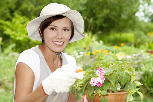 A woman wearing a white wide-brimmed hat, white gardening gloves, and a light grey t-shirt is tending to a potted flowering plant with pink blooms in a lush garden setting. The garden features a well-maintained lawn area with dense green foliage and various flowering plants in the background, including some yellow flowers. The scene is brightly lit, suggesting a sunny day, with natural sunlight illuminating the vibrant greenery and colorful flowers. The woman appears to be carefully inspecting or pruning the plant, indicating active gardening or outdoor maintenance work typical of professional landscaping or garden care services in Leyton. The environment shows a healthy, landscaped outdoor space with a mix of cultivated garden beds and natural elements, ideal for enhancing outdoor aesthetics and gardening efforts.