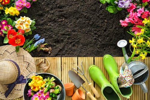 The image shows a section of a garden with freshly turned dark soil in the background, bordered by a wooden decking area. Along the edge of the decking, there are flowering plants with colorful blooms, including pink, purple, yellow, and red flowers, which appear to be planted in containers or directly in the soil. On the decking, there are gardening tools such as a trowel, a hand fork, and a small watering can, alongside a pair of bright green rain boots and a sunflower straw hat, indicating gardening activity. Additionally, there is a bowl filled with flowering plants and a polka-dotted gardening glove resting inside a watering can. The overall scene suggests preparations for planting or garden maintenance, with natural light illuminating the space, hinting at a clear day in Leyton or nearby in East London, aligning with gardening and landscaping services offered by Landscapers Leyton.