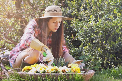 A young woman with long dark hair wearing a straw hat, plaid shirt, and gardening gloves is kneeling in a lush backyard garden, carefully tending to a flower bed filled with vibrant yellow and white blooms. The garden features a well-maintained green lawn in the foreground, with dense shrubbery and trees providing a natural backdrop. The sunlight filters through the leaves, casting a warm glow over the scene, and the woman appears focused on her gardening task, illustrating the outdoor care and landscaping services provided by Landscapers Leyton in the Leyton area, with the garden demonstrating a neat and inviting outdoor space suitable for relaxation and outdoor activities.